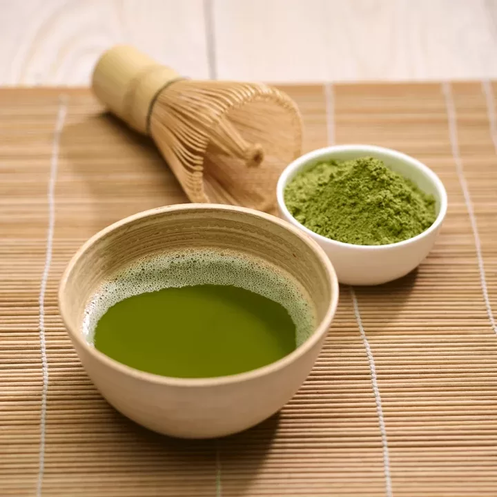 Chá verde matcha numa chavena de madeira, com pó de matcha numa chavena branca e um mexedor, em cima de um individual de mesa